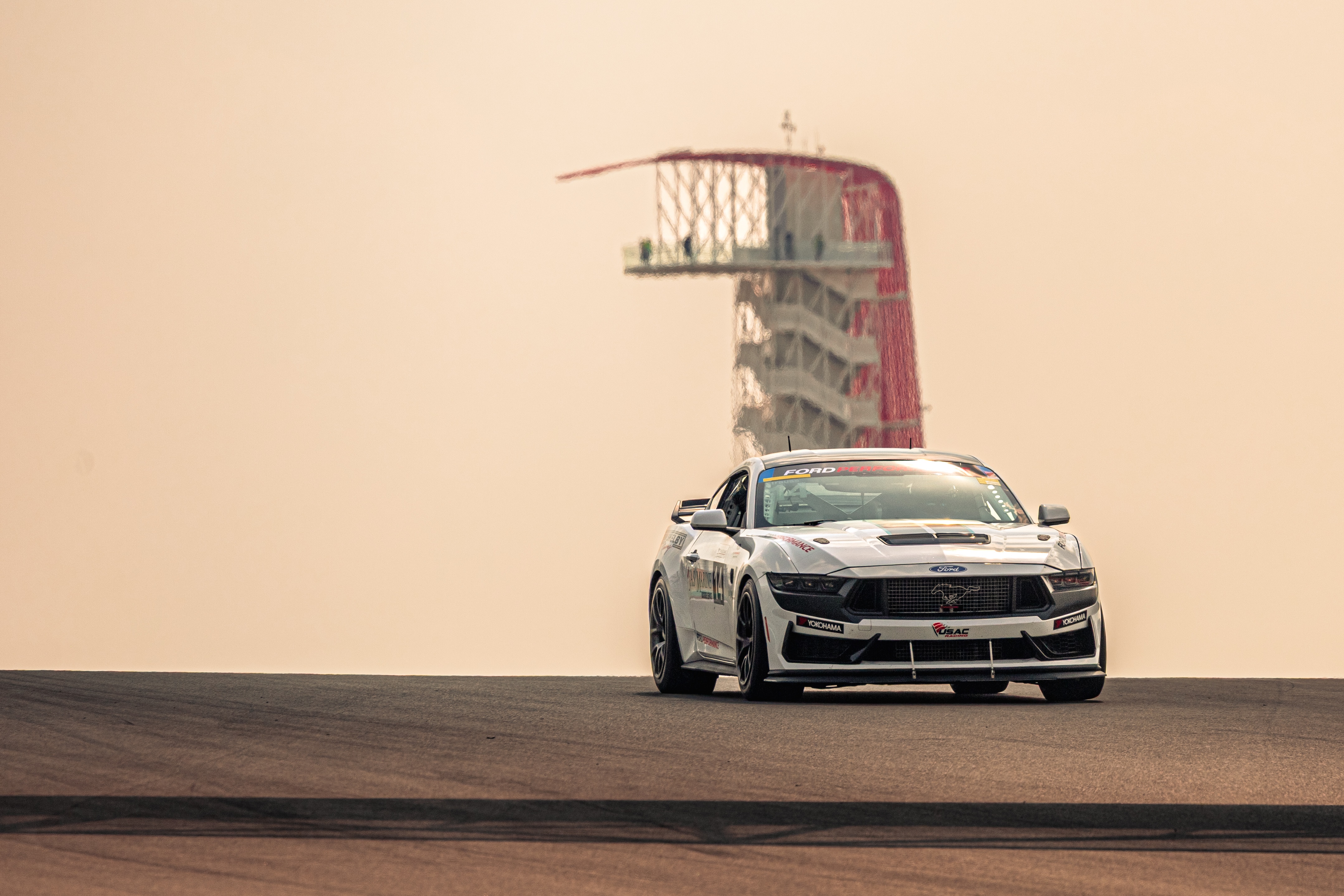 A white Mustang is parked on pavement. A tall structure with an observation deck at the top can be seen in the background.